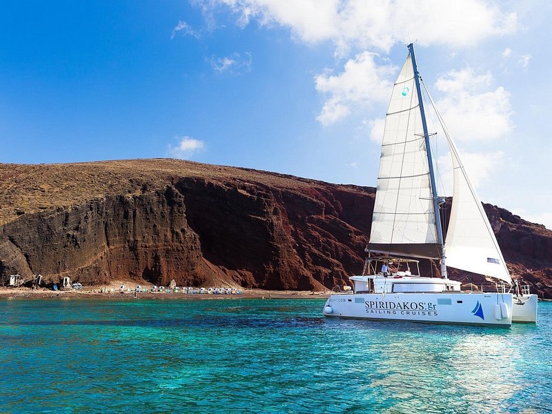 A catamaran moored in turquoise water under Santorini's red cliff