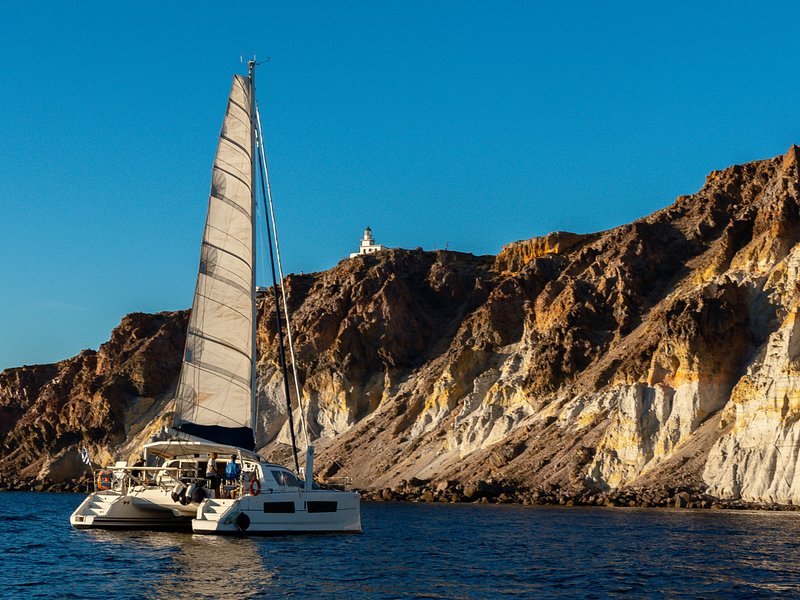A catamaran under sail below Santorini's white pumice cliffs
